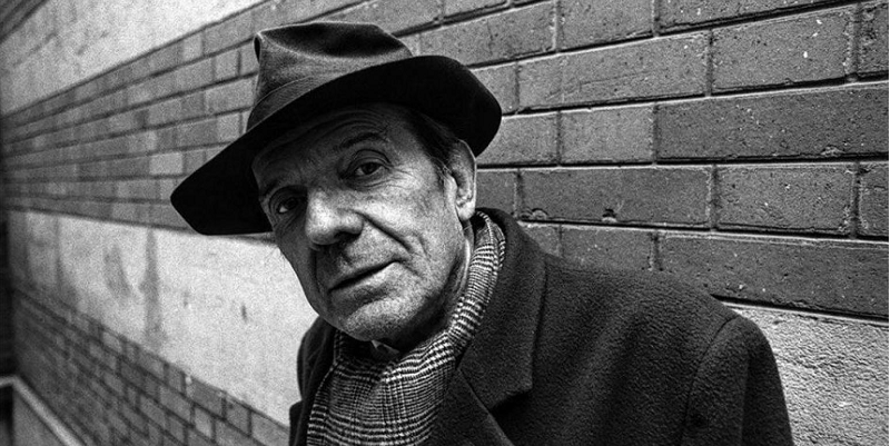 A black and white photo of Gilles Deleuze looking at the camera. He is wearing a hat. Behind him there is a brick wall.
